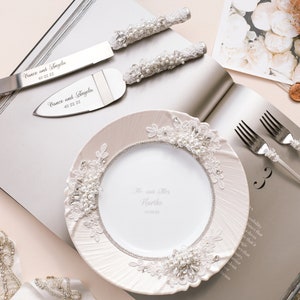 May include: A white cake plate with pearl and lace trim, a set of silver cake knives with pearl handles, and two silver forks with pearl accents. The knives are engraved with the names "Vince and Angela" and the date "03.03.23".