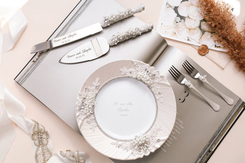 May include: A white cake plate with pearl and lace trim, a set of silver cake knives with pearl handles, and two silver forks with pearl accents. The knives are engraved with the names "Vince and Angela" and the date "03.03.23".