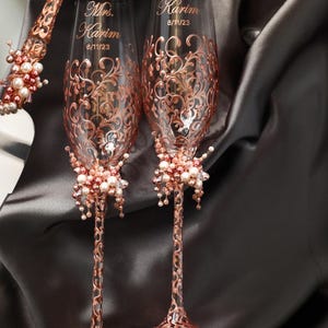 May include: Two rose gold wedding champagne flutes with ornate floral designs and the names "Mr. Karim" and "Mrs. Karim" with the date 6/11/23. Each flute has a cluster of pearls and crystals at the base.