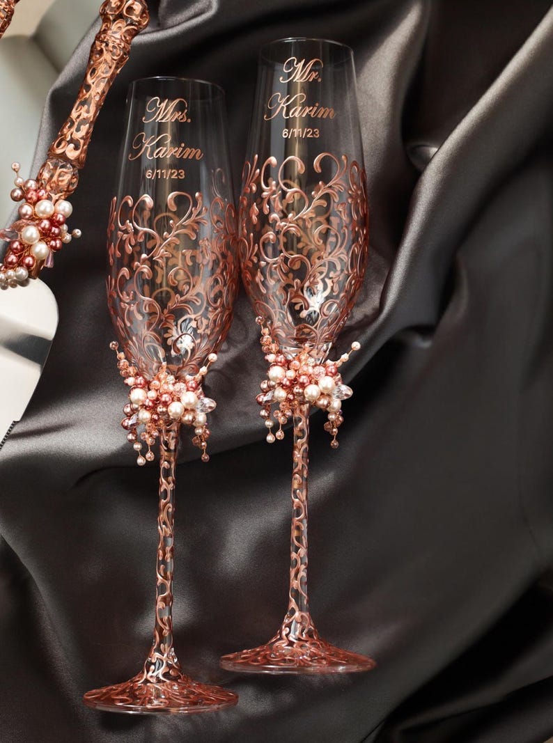 May include: Two rose gold wedding champagne flutes with ornate floral designs and the names "Mr. Karim" and "Mrs. Karim" with the date 6/11/23. Each flute has a cluster of pearls and crystals at the base.