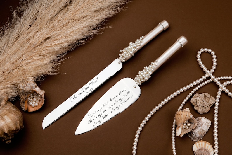 Puede incluir: Un juego de cuchillo y servidor de pastel de bodas plateado con detalles de perlas. El cuchillo est&aacute; grabado con "Mr. and Mrs. [Name] 09.26.2006". El servidor tiene una cita sobre el amor. Tambi&eacute;n hay perlas y conchas decorativas.