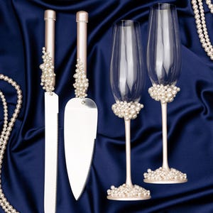 Puede incluir: Un juego de cuchillo y servidor para pastel de bodas con detalles de perlas, junto con dos copas de champ&aacute;n con detalles de perlas. Los art&iacute;culos se presentan sobre un fondo de tela de sat&eacute;n azul oscuro, con collares de perlas.