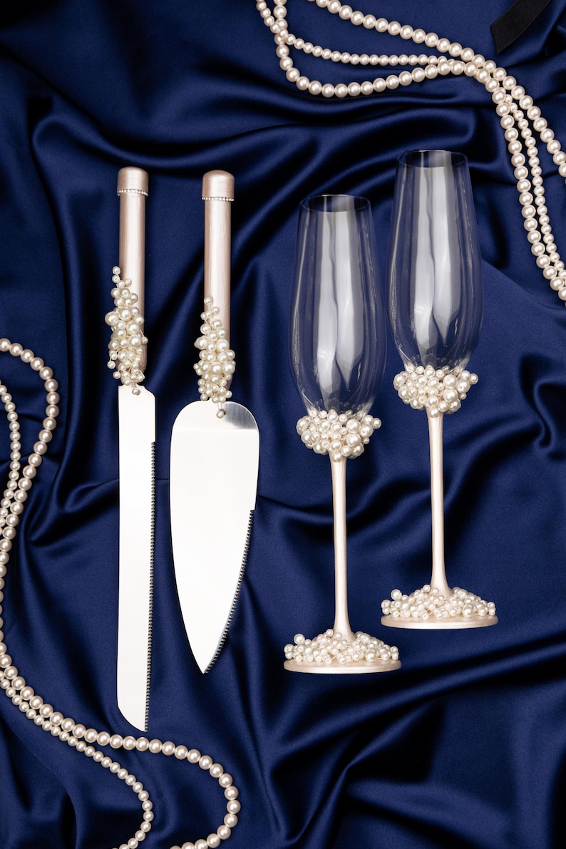 Puede incluir: Un juego de cuchillo y servidor para pastel de bodas con detalles de perlas, junto con dos copas de champ&aacute;n con detalles de perlas. Los art&iacute;culos se presentan sobre un fondo de tela de sat&eacute;n azul oscuro, con collares de perlas.