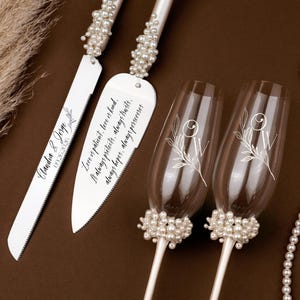 May include: A wedding cake knife and server set with pearl accents and engraved text. Two champagne flutes with floral designs and pearl bases are also present. A pearl necklace and decorative elements complete the arrangement.