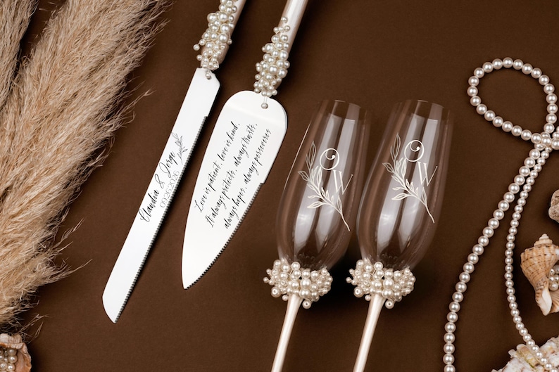 May include: A wedding cake knife and server set with pearl accents and engraved text. Two champagne flutes with floral designs and pearl bases are also present. A pearl necklace and decorative elements complete the arrangement.