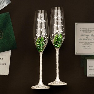 May include: Two clear glass champagne flutes with silver-toned stems and bases, featuring white scrollwork designs, green and white pearl-like embellishments, and the etched text MR. Hollar 5/23/26 and MRS. Hollar 5/23/26, accompanied by wedding stationery.