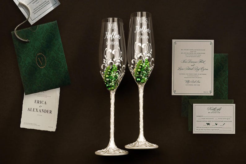 May include: Two clear glass champagne flutes with silver-toned stems and bases, featuring white scrollwork designs, green and white pearl-like embellishments, and the etched text MR. Hollar 5/23/26 and MRS. Hollar 5/23/26, accompanied by wedding stationery.