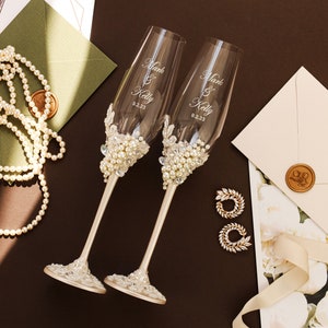 May include: Two clear glass champagne flutes featuring white pearl and clear crystal embellishments on the stems and bases. The glass bowls are etched with the text Mark & Kelly and the date 9.2.23. The stems are finished in a smooth champagne gold tone.