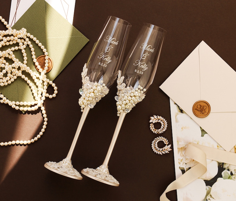 May include: Two clear glass champagne flutes featuring white pearl and clear crystal embellishments on the stems and bases. The glass bowls are etched with the text Mark & Kelly and the date 9.2.23. The stems are finished in a smooth champagne gold tone.