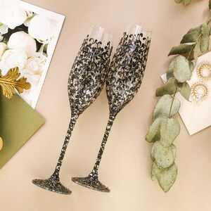 May include: Two champagne flutes with black and white floral designs. The flutes are on a beige surface with a green envelope and a sprig of eucalyptus.