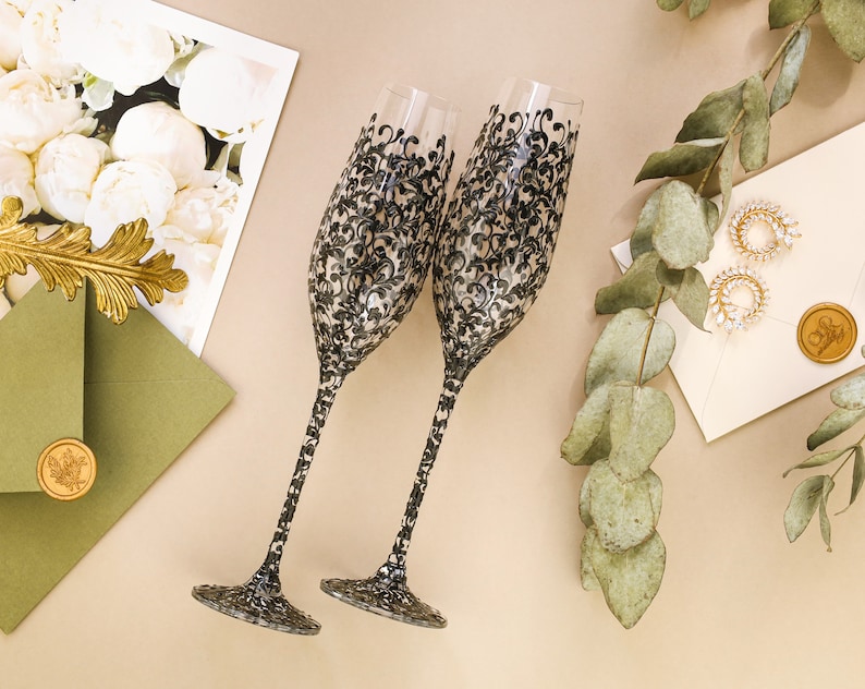 May include: Two champagne flutes with black and white floral designs. The flutes are on a beige surface with a green envelope and a sprig of eucalyptus.