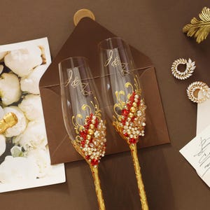 May include: Two clear glass champagne flutes with gold stems, featuring red and white crystal embellishments and the engraved initials J&K. The set is displayed alongside a floral card, a brown envelope, and a handwritten note.