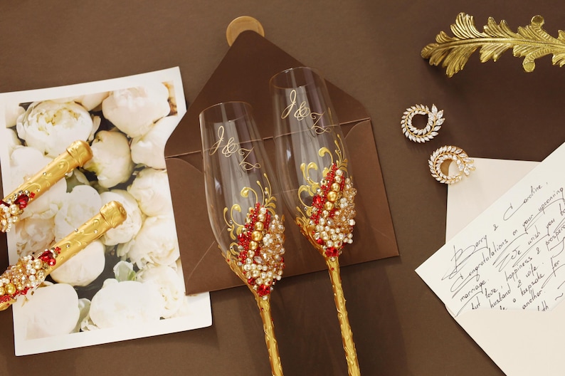 May include: Two clear glass champagne flutes with gold stems, featuring red and white crystal embellishments and the engraved initials J&K. The set is displayed alongside a floral card, a brown envelope, and a handwritten note.
