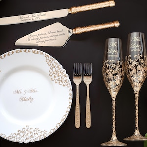May include: A collection of wedding accessories, including a cake knife and server, a decorative plate, two champagne flutes, and two forks. The items are gold and white, with ornate designs and personalized text. The plate reads "Mr. & Mrs. Shultz."