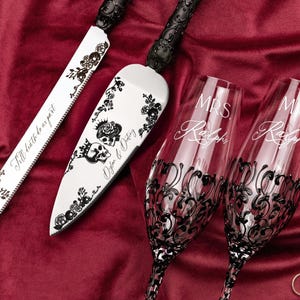 May include: A cake knife and server set with black ornate handles and silver blades featuring black skull and floral graphics, plus two clear champagne flutes with black filigree bases and white text reading MRS. Ralphs and MR. Ralphs.