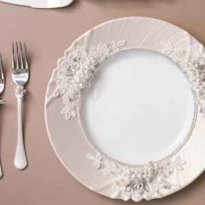 May include: A white ceramic plate with a decorative lace trim and pearl accents. Two silver forks with the names "Angela" and "Vince" engraved on them are placed on a beige surface.