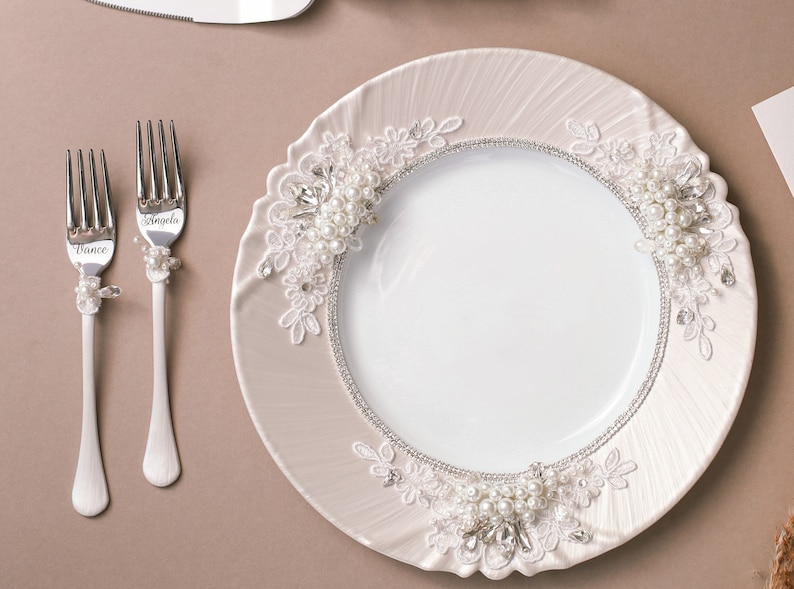 May include: A white ceramic plate with a decorative lace trim and pearl accents. Two silver forks with the names "Angela" and "Vince" engraved on them are placed on a beige surface.