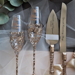 Könnte beinhalten: Ein Hochzeitstorten-Schneideset mit zwei Champagnerflöten. Die Flöten sind mit goldenen Filigranen und den Worten "Mr. Wilson 10.20.2022" und "Mrs. Wilson 10.20.2022" verziert. Das Tortenmesser trägt die Worte "Samantha und Raymond 10.20.2022" und "You will Forever be my Always" eingraviert.