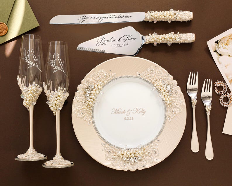 May include: Elegant wedding set featuring a cake knife and server, a plate, two champagne flutes, and two forks. The knife and server have pearl and crystal accents. The plate and flutes are decorated with floral designs and the text "You are my greatest adventure".