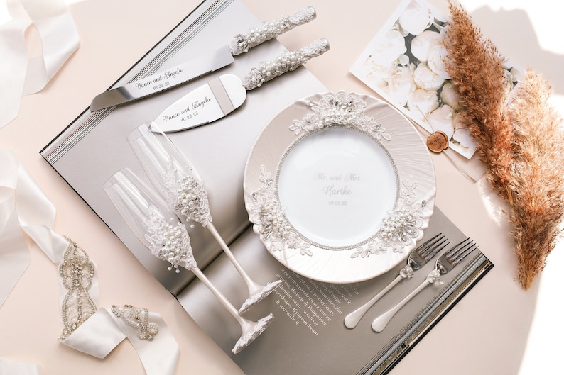May include: A white ceramic plate with a lace design and pearl accents. The plate has the text "Mr. and Mrs. Hardie" and a date. Two champagne flutes with pearl and lace accents, a silver cake knife and server, and a white ribbon are also pictured.