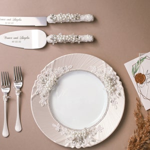 May include: A white ceramic plate with a lace design and pearl accents. Two silver forks with pearl accents and two silver cake knives with pearl accents and the text "Vince and Angela" are on a brown surface.