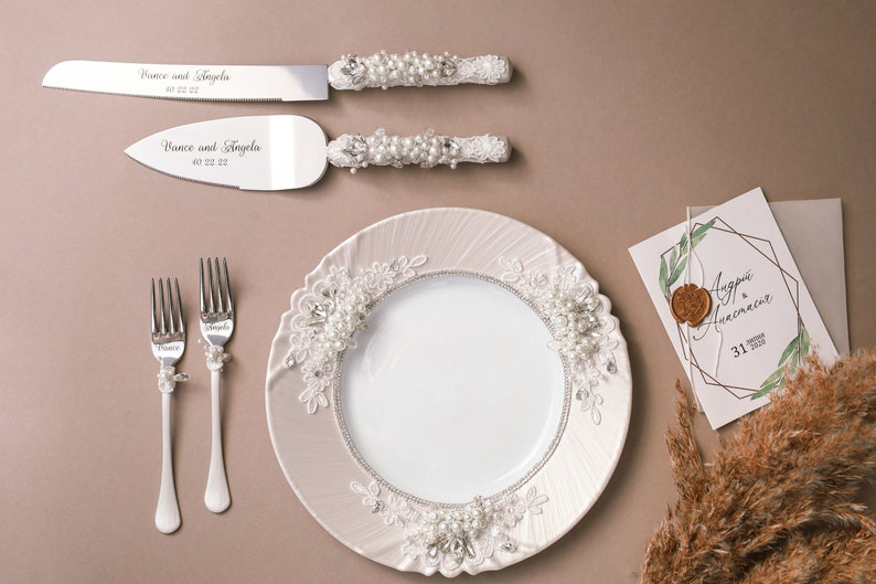 May include: A white ceramic plate with a lace design and pearl accents. Two silver forks with pearl accents and two silver cake knives with pearl accents and the text "Vince and Angela" are on a brown surface.