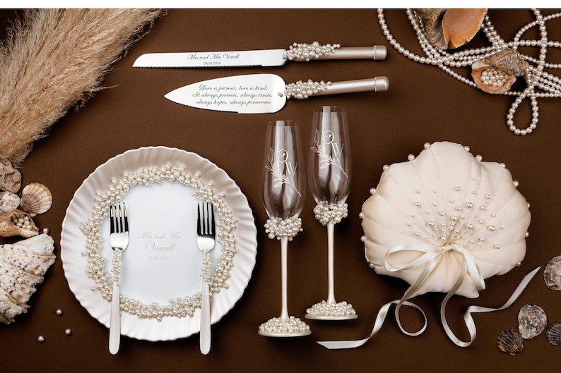 May include: Wedding table setting with a cake knife and server, two champagne flutes, a ring pillow, and a plate with forks. The items are decorated with pearls and sit on a brown surface. The knife and server have text.
