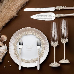 May include: Elegant wedding set featuring a cake knife and server, a plate with two forks, and two champagne flutes. The set is adorned with pearls and includes engraved text. The plate and flutes have floral designs.