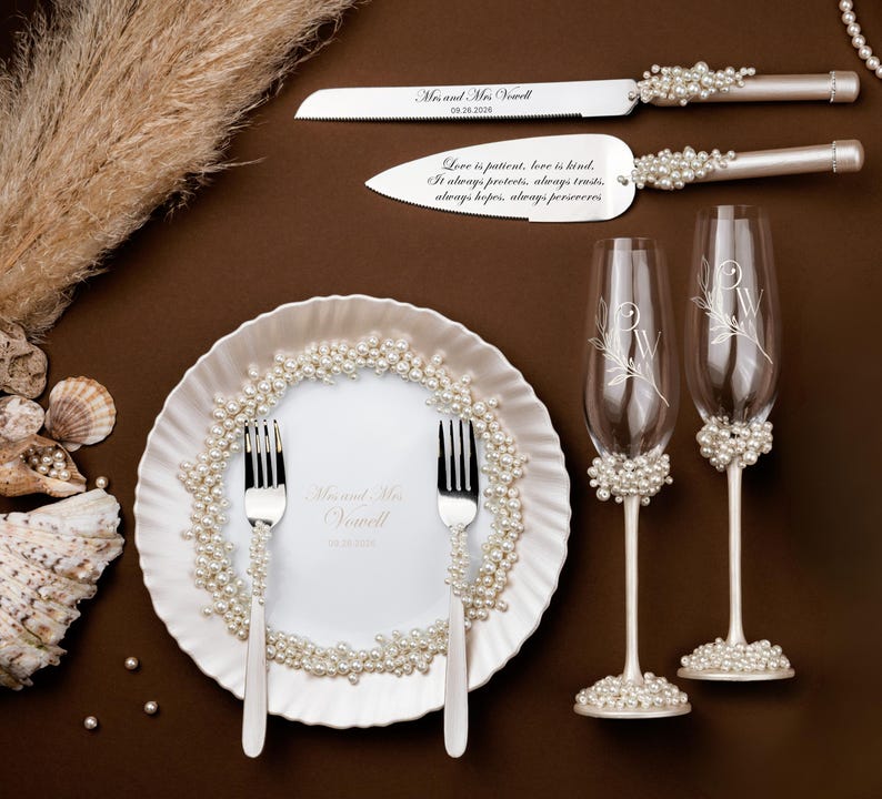 May include: Elegant wedding set featuring a cake knife and server, a plate with two forks, and two champagne flutes. The set is adorned with pearls and includes engraved text. The plate and flutes have floral designs.