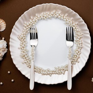 Puede incluir: Un plato blanco con borde ondulado, adornado con perlas y dos tenedores plateados. Los tenedores est&aacute;n decorados con perlas. El plato est&aacute; sobre un fondo marr&oacute;n, lo que sugiere un tema de comedor o decoraci&oacute;n.