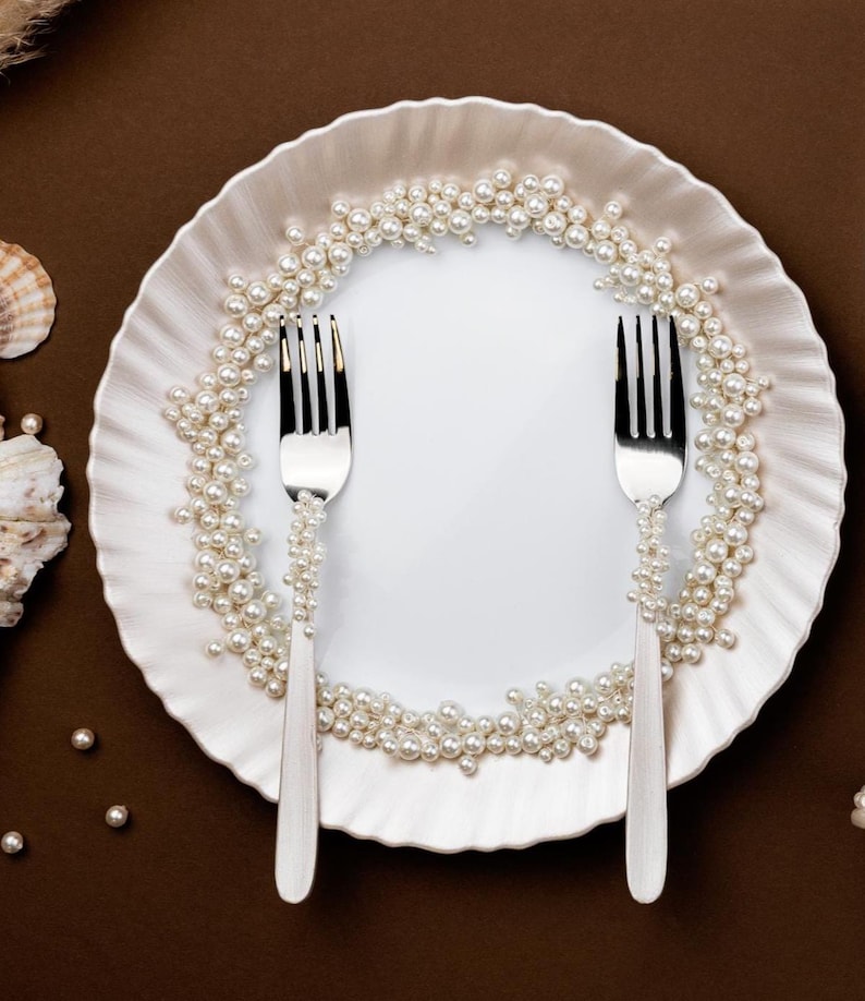 Puede incluir: Un plato blanco con borde ondulado, adornado con perlas y dos tenedores plateados. Los tenedores est&aacute;n decorados con perlas. El plato est&aacute; sobre un fondo marr&oacute;n, lo que sugiere un tema de comedor o decoraci&oacute;n.