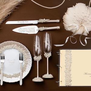 Puede incluir: Accesorios de boda dispuestos sobre una superficie marr&oacute;n. Incluye un cuchillo y una esp&aacute;tula para pastel, dos copas de champ&aacute;n, una almohada para anillos, un libro de visitas y un plato con tenedores. Todos los art&iacute;culos est&aacute;n decorados con perlas. El servidor de pastel tiene el texto "Love is patient, love is kind."