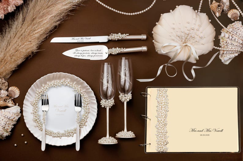 Puede incluir: Accesorios de boda dispuestos sobre una superficie marr&oacute;n. Incluye un cuchillo y una esp&aacute;tula para pastel, dos copas de champ&aacute;n, una almohada para anillos, un libro de visitas y un plato con tenedores. Todos los art&iacute;culos est&aacute;n decorados con perlas. El servidor de pastel tiene el texto "Love is patient, love is kind."