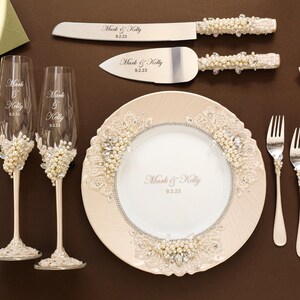 Puede incluir: Un conjunto de boda que incluye dos copas de champ&aacute;n, un cuchillo para pastel, un servidor de pastel y dos tenedores, decorados con perlas blancas, encaje y cristales transparentes. Cada pieza est&aacute; grabada con el texto Mark & Kelly y la fecha 9.2.23. Se incluye un plato a juego.