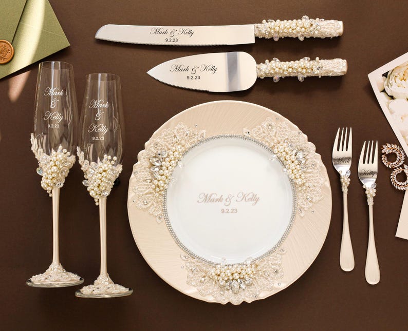 Puede incluir: Un conjunto de boda que incluye dos copas de champ&aacute;n, un cuchillo para pastel, un servidor de pastel y dos tenedores, decorados con perlas blancas, encaje y cristales transparentes. Cada pieza est&aacute; grabada con el texto Mark & Kelly y la fecha 9.2.23. Se incluye un plato a juego.