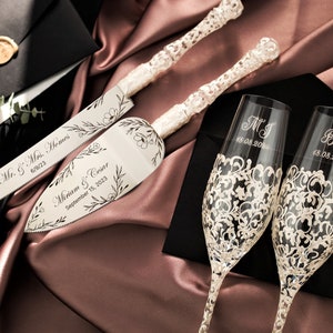 May include: A wedding cake cutting set with two silver knives and two champagne flutes. The knives are engraved with the names "Mr. & Mrs. Homes" and "Miriam & Cesar" with their wedding date. The champagne flutes are engraved with the names "MJ" and "Ben" with their wedding dates.