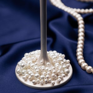 May include: A decorative wine glass with a white base and stem, adorned with numerous small, round pearls. A strand of pearls rests on a deep blue satin surface, complementing the elegant design. The pearls are of varying sizes.
