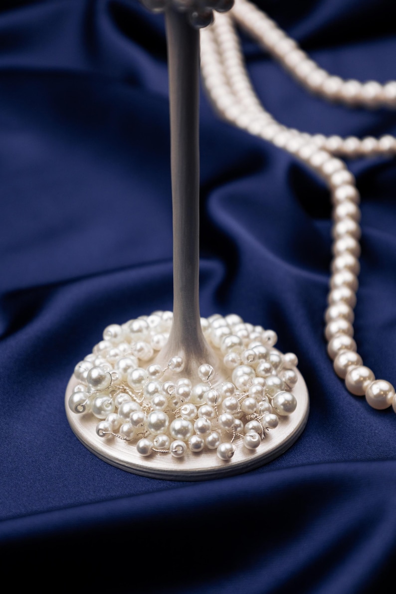 May include: A decorative wine glass with a white base and stem, adorned with numerous small, round pearls. A strand of pearls rests on a deep blue satin surface, complementing the elegant design. The pearls are of varying sizes.