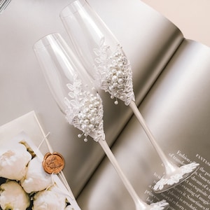 May include: Two champagne flutes with white lace and pearl embellishments. The flutes are on a white surface with a book and a bouquet of white flowers in the background.