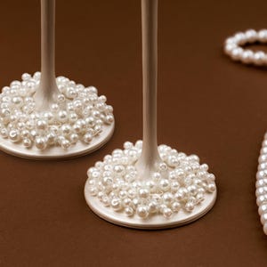 May include: Two champagne glasses with pearl-covered bases and long stems. A strand of pearls is draped on a brown surface. The glasses have a classic, elegant design, perfect for special occasions.