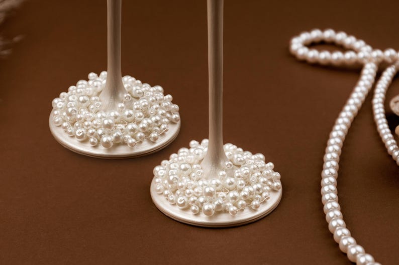 May include: Two champagne glasses with pearl-covered bases and long stems. A strand of pearls is draped on a brown surface. The glasses have a classic, elegant design, perfect for special occasions.
