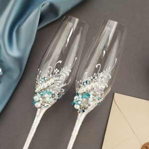 May include: Two clear glass champagne flutes featuring white painted floral flourishes and an embellishment of blue, clear, and white beads and pearls clustered at the base of the bowl. The stems are wrapped in a white material with small clear crystals.