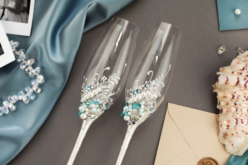 May include: Two clear glass champagne flutes featuring white painted floral flourishes and an embellishment of blue, clear, and white beads and pearls clustered at the base of the bowl. The stems are wrapped in a white material with small clear crystals.