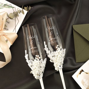 May include: Two clear glass champagne flutes with a white lace and pearl embellishment. The flutes are engraved with the names "Khaled & Qamar" and the date "08.25.2023".