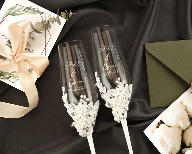 May include: Two clear glass champagne flutes with a white lace and pearl embellishment. The flutes are engraved with the names "Khaled & Qamar" and the date "08.25.2023".
