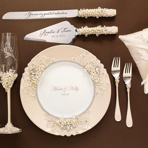 May include: A wedding set featuring two champagne flutes, a cake knife and server, a plate, two forks, and a ring pillow. The flutes, knife, and server have pearl and lace accents. The plate is engraved with Mark & Kelly 9.2.23 and lace trim.