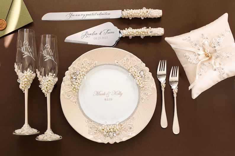 May include: A wedding set featuring two champagne flutes, a cake knife and server, a plate, two forks, and a ring pillow. The flutes, knife, and server have pearl and lace accents. The plate is engraved with Mark & Kelly 9.2.23 and lace trim.