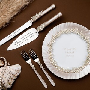 Puede incluir: Un juego para servir pastel de bodas con detalles de perlas, que incluye un cuchillo y un servidor, junto con dos tenedores y un plato decorativo. El juego est&aacute; sobre una superficie marr&oacute;n con texto.