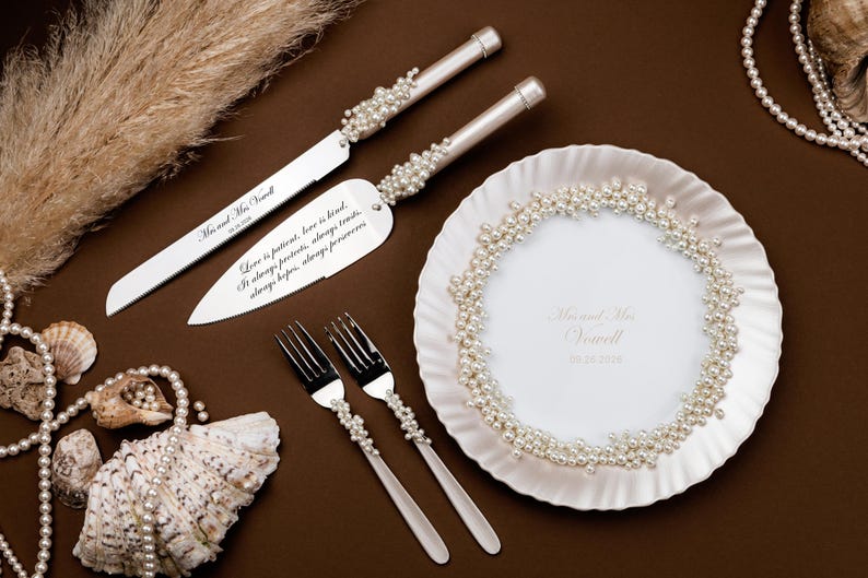 Puede incluir: Un juego para servir pastel de bodas con detalles de perlas, que incluye un cuchillo y un servidor, junto con dos tenedores y un plato decorativo. El juego est&aacute; sobre una superficie marr&oacute;n con texto.