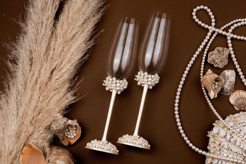 Puede incluir: Dos copas de champ&aacute;n con detalles de perlas en la base y el tallo, sobre un fondo marr&oacute;n. Perlas decorativas y conchas marinas dispuestas cerca. Hierba de la Pampa seca a&ntilde;ade un elemento natural a la composici&oacute;n.